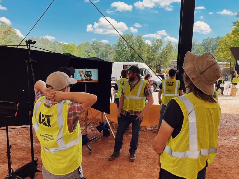 A Caterpillar crew in high-visibility safety vests on an outdoor filming location, discussing setup in front of a monitor displaying heavy machinery.