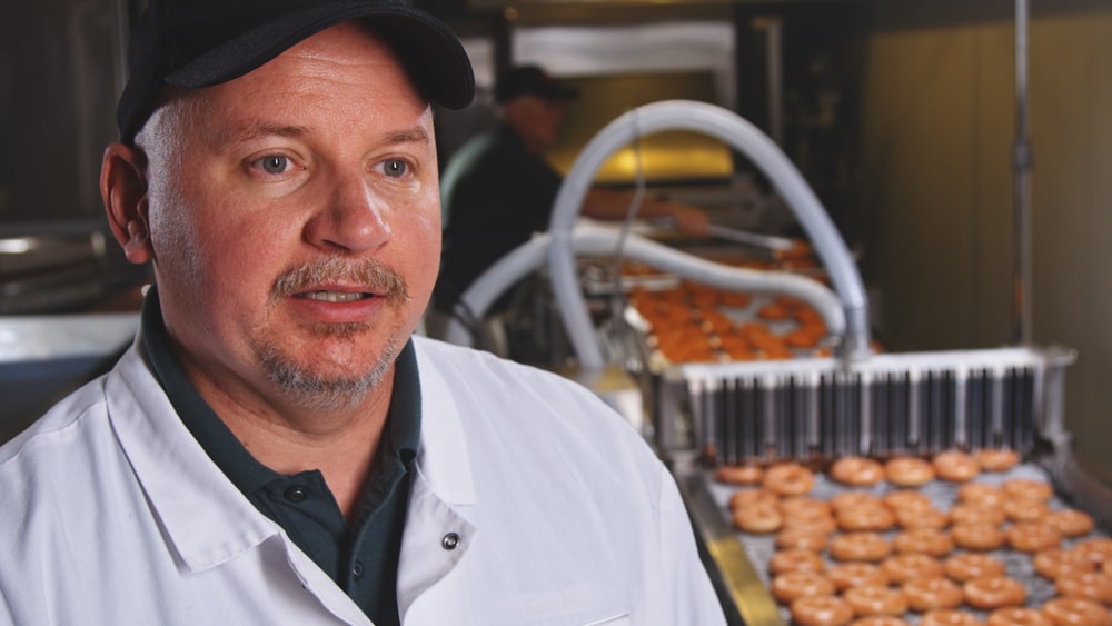 Krispy Kreme bakery manager in a white coat stands near a conveyor belt filled with freshly made donuts, overseeing quality control in the production line.