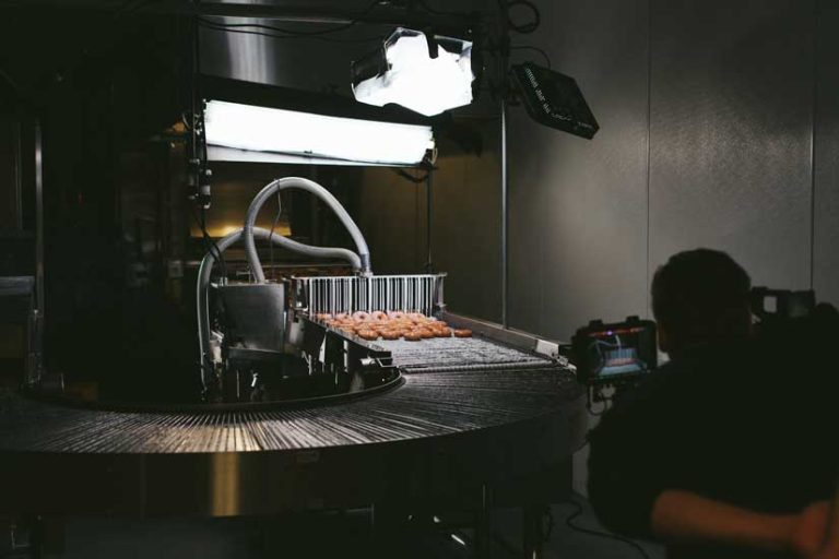 Krispy Kreme donuts moving along a production conveyor belt under professional lighting, with a cameraman capturing the process.