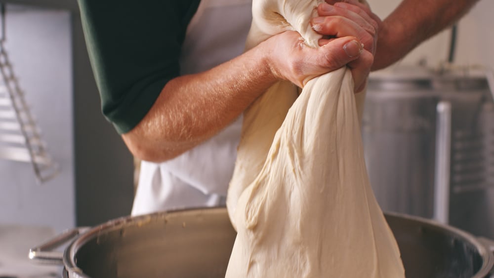 Close-up of a baker's hands stretching fresh dough in a metal bowl, showcasing the craftsmanship and care in Krispy Kreme's dough preparation process.