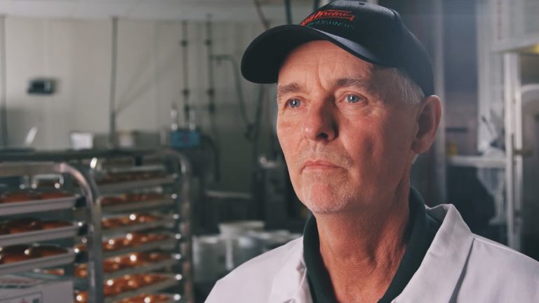 Krispy Kreme production supervisor in a white coat and branded cap stands in front of trays of freshly made donuts, overseeing the quality and efficiency of the production process.