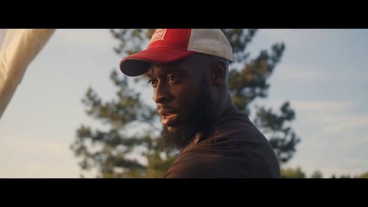 A farmer at Sankofa Farms in a red and white cap, gazing intently with a look of dedication, against a backdrop of trees and sky.