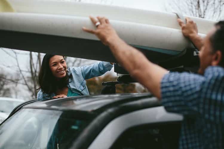 A smiling woman and a man loading a surfboard onto the roof of an SUV, preparing for an outdoor adventure, highlighting South State Bank’s role in helping families plan for memorable trips and experiences.
