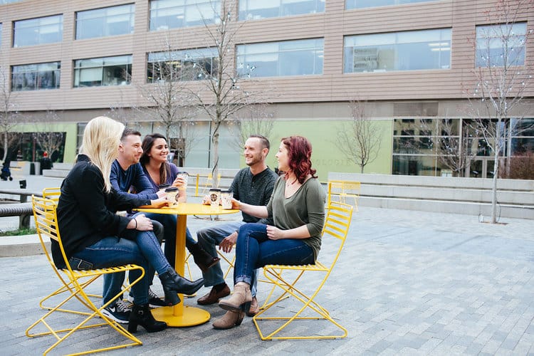 A group of five people sitting at a yellow table outside a modern building, enjoying coffee and casual conversation, representing South State Bank’s approach to building strong relationships with its customers.